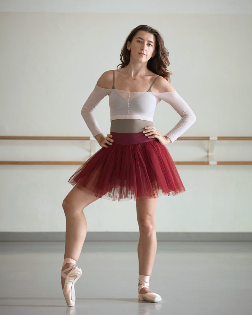 Taylor Naturkas, wearing a burgundy practice tutu, brown leotard and pink shrug, faces the camera and stands with her right foot propped onto pointe and her hands on her hips. She looks at the camera confidently.