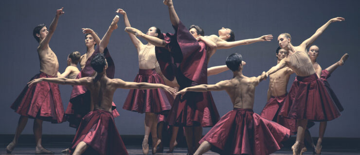 Twelve dancers from the Paris Opéra Junior Ballet create a tableau onstage with various poses. One dancer is lifted up on her back, her top leg pointing upward. Other dancers lunge forward, tendu, balance in sous-sus, and pose in arabesque. They all wear shiny, full, deep-red skirts and either nothing or a nude-colored leotard on top.