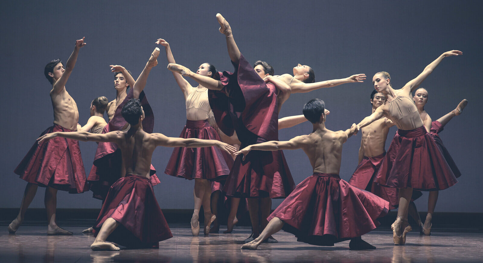 Twelve dancers from the Paris Opéra Junior Ballet create a tableau onstage with various poses. One dancer is lifted up on her back, her top leg pointing upward. Other dancers lunge forward, tendu, balance in sous-sus, and pose in arabesque. They all wear shiny, full, deep-red skirts and either nothing or a nude-colored leotard on top.