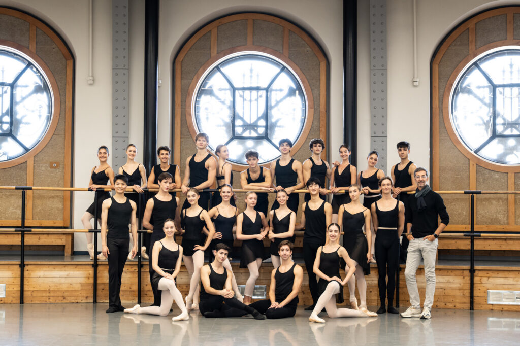 Paris Opéra Junior Ballet—24 dancers and one director—pose for a group photo arranged in three lines. They pose in front of three large circular windows in a bright ballet studio.