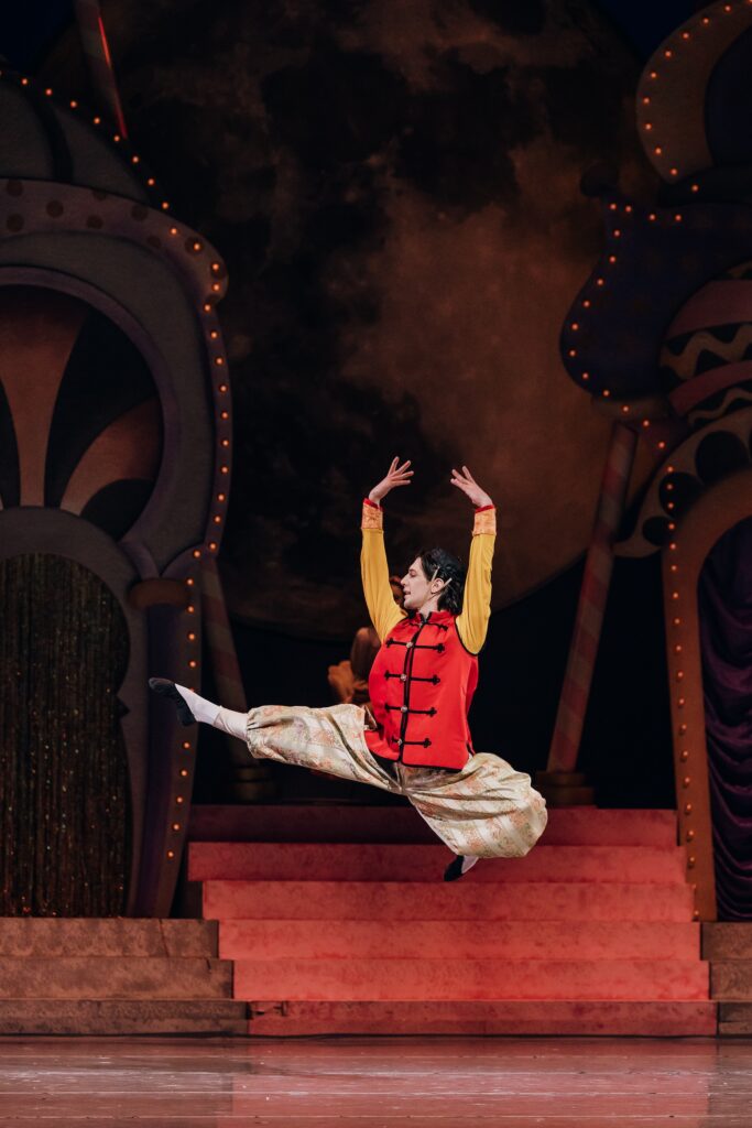 On stage, Andrew Sackett does an Italian pas de chat with both arms raised overhead. He is dressed in a red-and-gold top, gold pants with red designs, and black flat shoes.