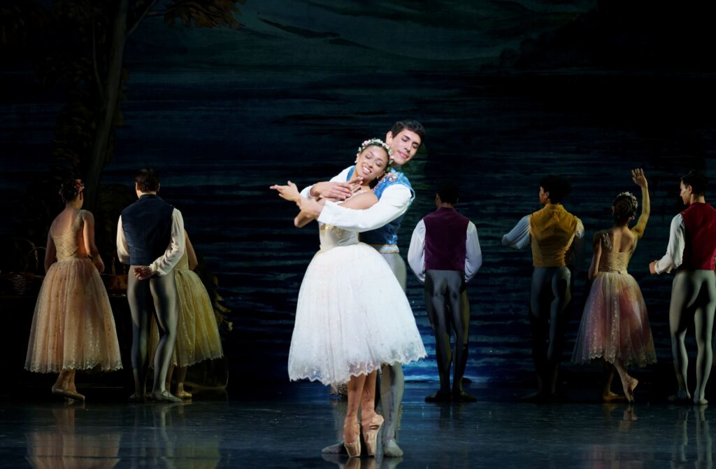 Onstage, wearing a white romantic tutu and floral head piece, Rachel Zinman stands in sous-sus en point. Her partner, Emanuel Talongoin, wears a blue vest, white tunic, and sage green tights, as he wraps his arms around her.