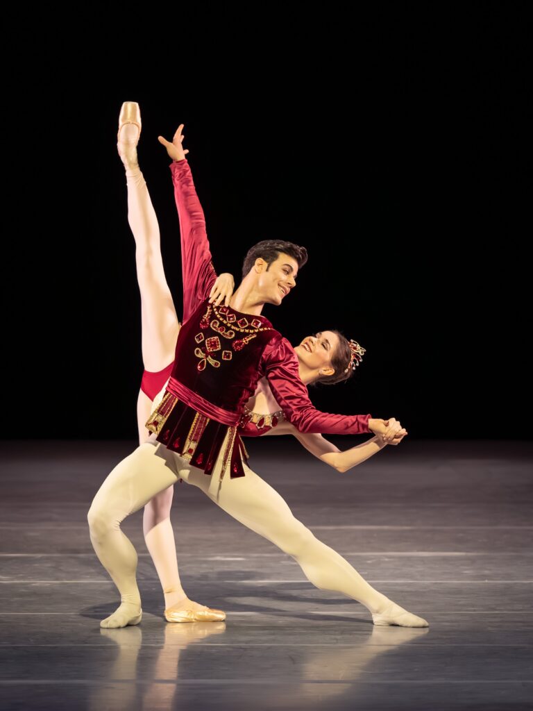 Trenary smiles sunnily at her partner as she pauses in an arabesque penché behind him, supporting herself with a hand on his shoulder and another in his proffered palm. Her partner lunges to the side with an arm extended high to parallel her working leg, and looks over his shoulder back at her.