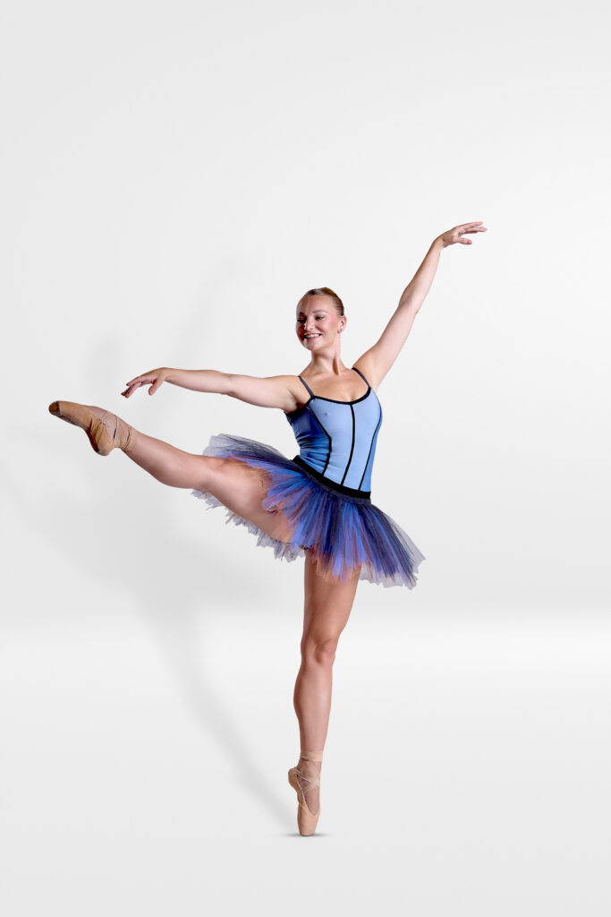 Whitney Hester, wearing a blue leotard and tutu, poses en point with her right let extended to the side. Her right arm is extended to the side, while her left is up.