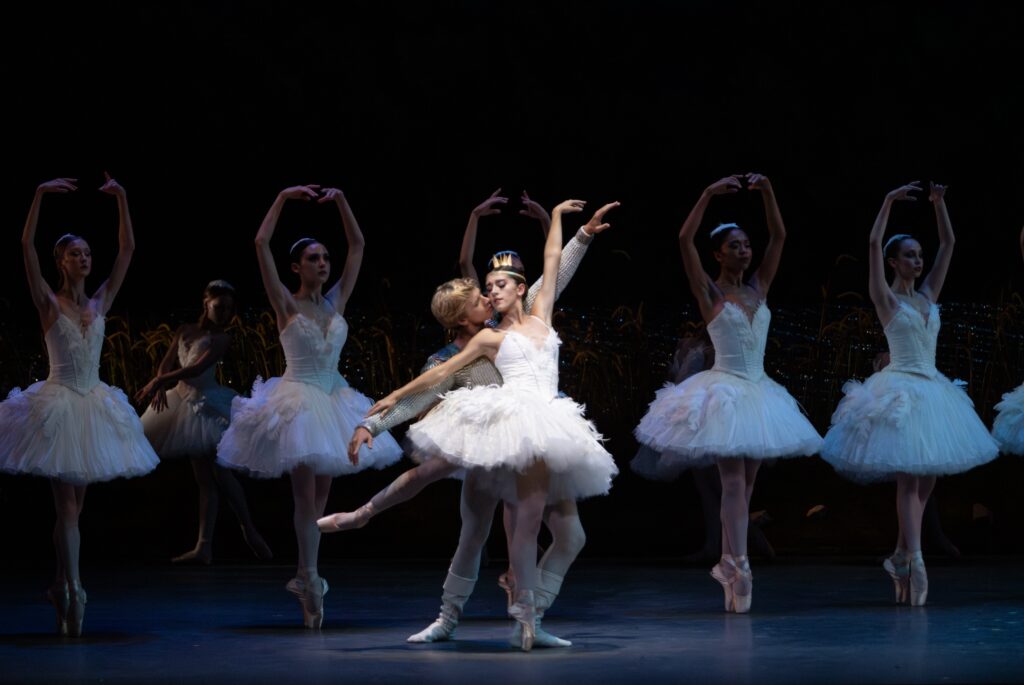 Taylor Naturkas, wearing a white tutu trimmed with feathers and a gold crown, does a low arabesque on pointe, leaning slightly on her partner's chest as he lunges behind her, their faces almost touching. A corps de ballet in white tutus balance in sus-sous on pointe behind them.