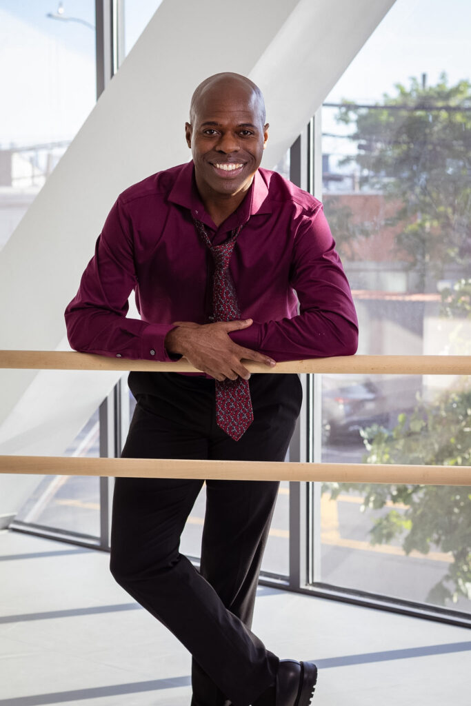 Christopher Charles McDaniel smiles for a portrait while leaning on a double ballet barre in a brightly lit studio.