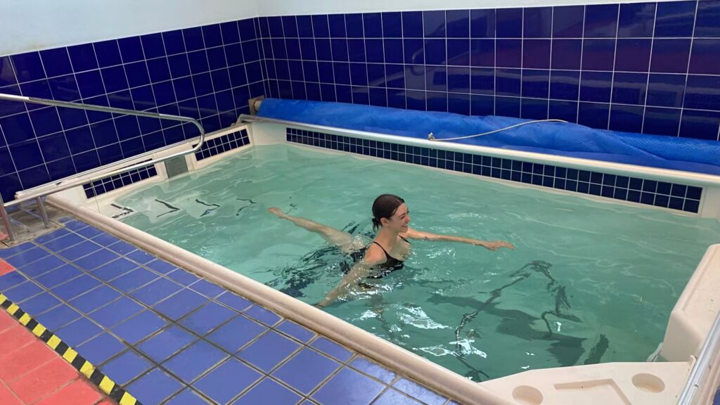 A dancer wearing a swimsuit practices an arabesque in a small, indoor pool. Royal blue tiles line the floor and walls of the facility.