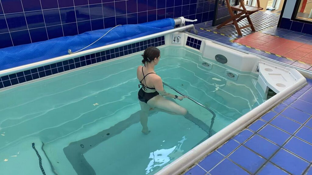 A woman in a black swimsuit is shown from behind posing in retiré relevé in a small indoor pool. She places a fnger on the pool's handrail for balance.