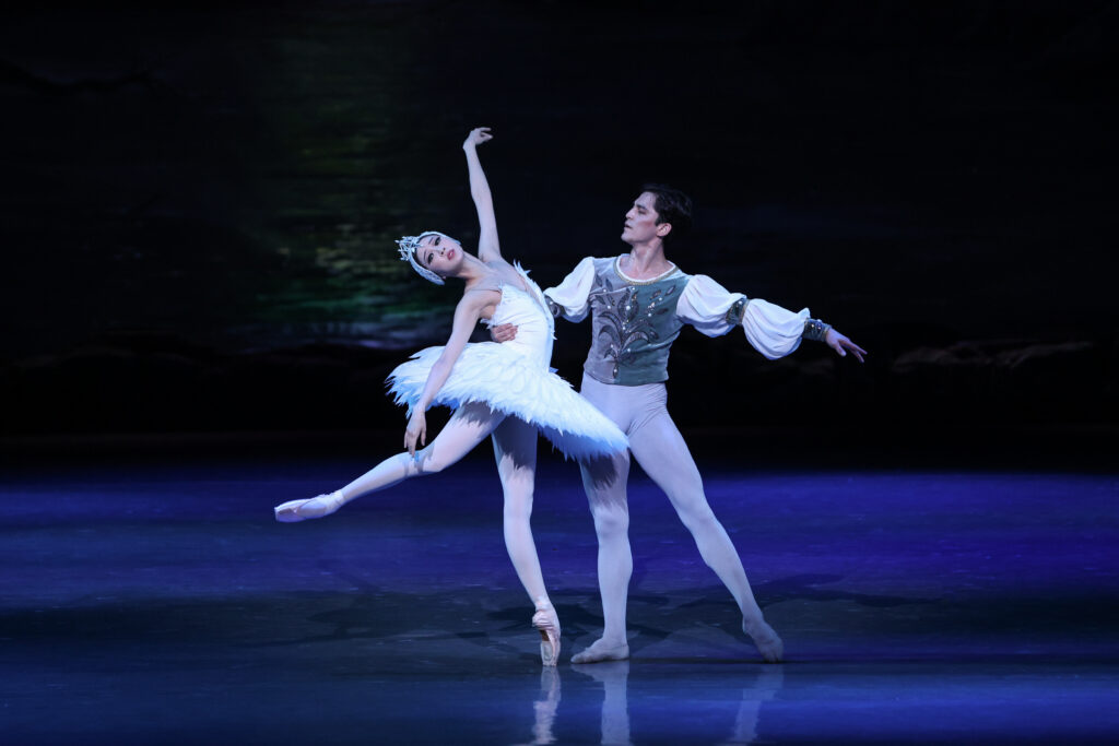 Onstage in dark bluish lighting, two dancers dressed as Odette and Prince Seigfried perform a pas de deux. Odette leans back in a low attitude derriere on pointe, arms extended long, as Siegfried counterbalances her with one arm wrapped around her waist as he poses in tendu.