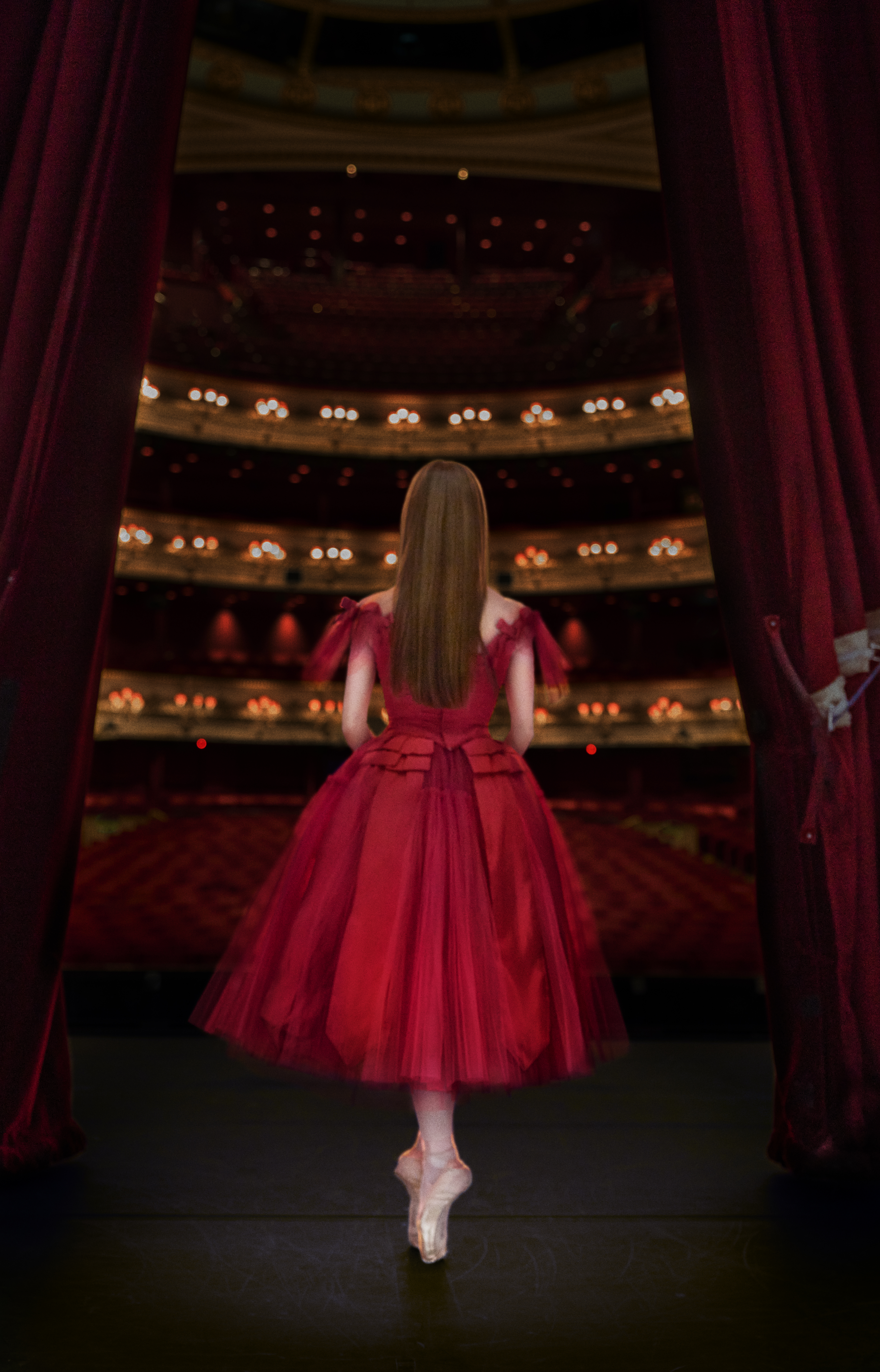 Marianela Nuñez, in a bright red, full dress with tulle and ribbons, stands in sous-sus on pointe facing away from the camera. The two red Royal Opera House curtains are partially drawn on either side of her, exposing the house behind her.