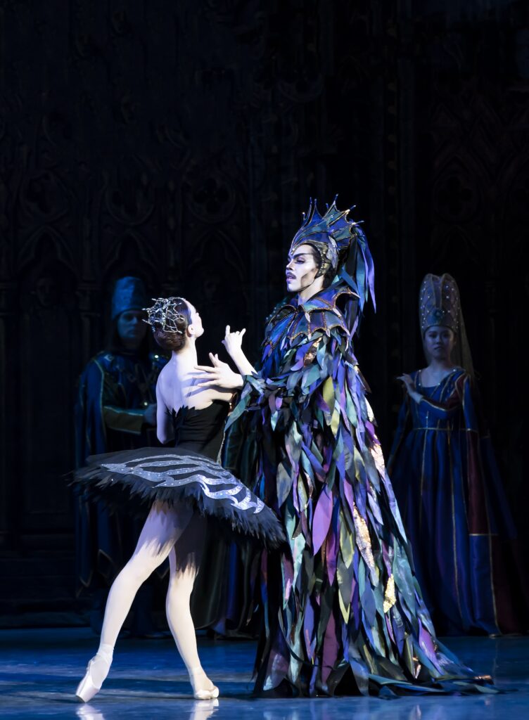 Onstage, two dancers dressed as Odile and Von Rothbart pretend to conspire. Odile stands in tendu derriere, lifting one arm with her elbow bent as she faces Rothbart. The sorcerer, in a lavish feathered costume, stands stoically as he looks into her eyes and places one hand on her shoulder.