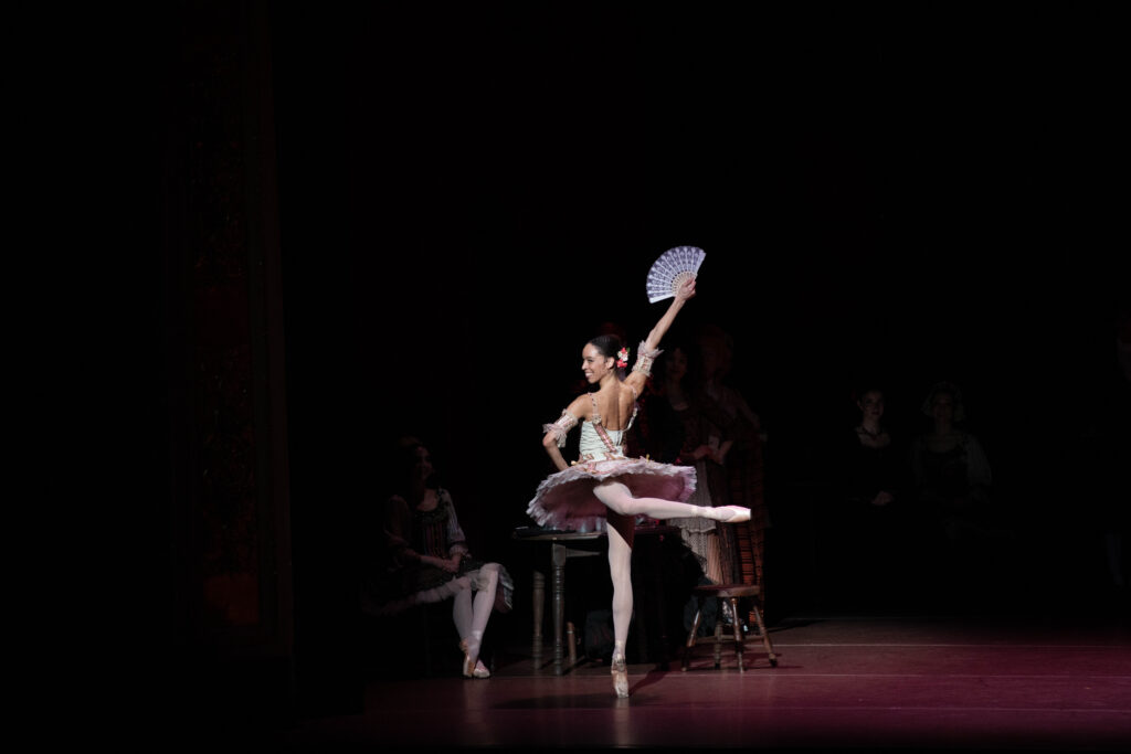 Chyrstyn Mariah Fentroy poses in piqué arabesque croisé derriere, her hips facing the back left corner onstage as she twists her upper body to look at the audience. She wears a white tutu with red and black accents, posing with one hand on her hip and the other raised as she flicks a fan open.