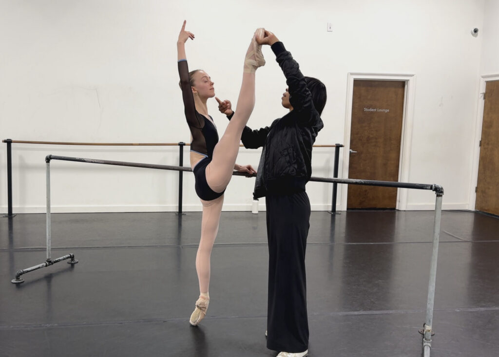 Georné Aucoin faces a young ballet student at the barre, holding the dancer's top foot in a high battement position. The student wears a long-sleeved black leotard, tights, and pointe sheos.