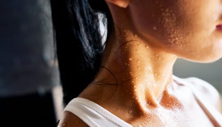 Close up of the shoulders and neck of a young woman sweating profusely. She wears a white leotard and her dark hair in a ponytail.