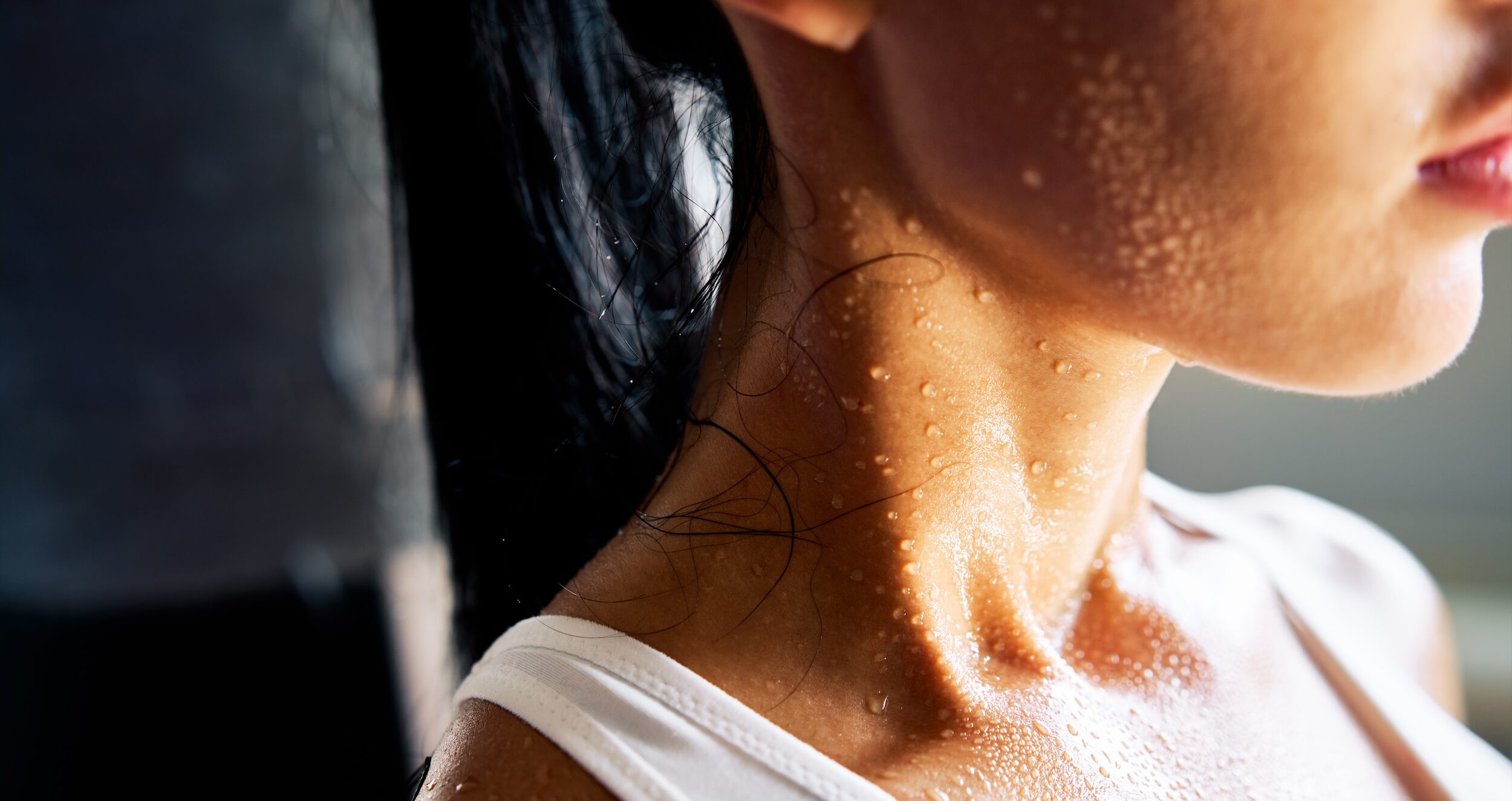 Close up of the shoulders and neck of a young woman sweating profusely. She wears a white leotard and her dark hair in a ponytail.