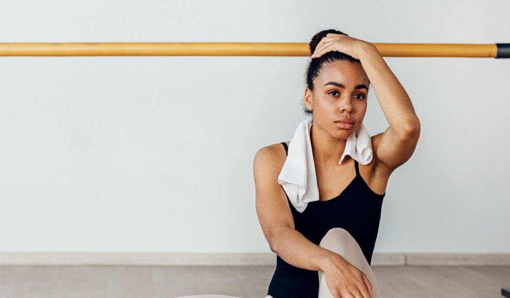 A young ballet dancer wearing a black leotard and pink tights sits on a floor and rests in a studio. She has a towel around her shoulders and rests her left hand on her head, looking exhausted.
