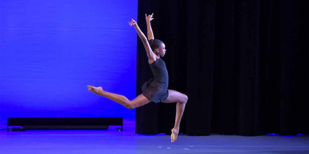 Onstage at a ballet competition, Nola Paulina jumps with both legs bent in parallel, one lifted front and one in back, with her arms reaching up. She faces the right and wears a short black dress.