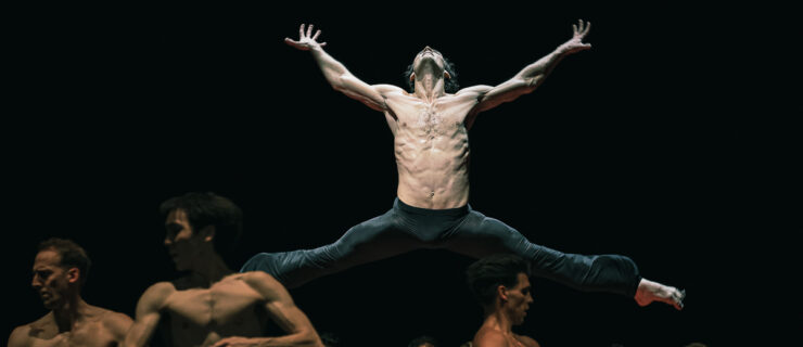 Hugo Marchand, surrounded by an ensemble of dancers, jumps with his legs split sideways and his arms stretched out to the sides. He tilts his head back and splays his fingers. He and the other dancers all wear black pants.