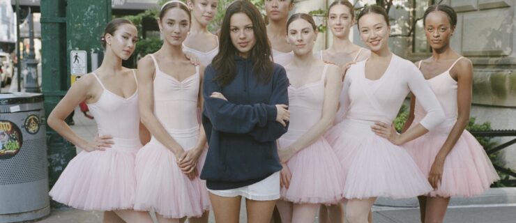 Olivia Rodrigo, a young woman wearing a large, blue, long-sleeved shirt, whort white skirt, and black knee-length boots, poses with her arms crossed with eight ballerinas. The dancers are all costumed in pink tutus. They pose outside on a New York City sidewalk.