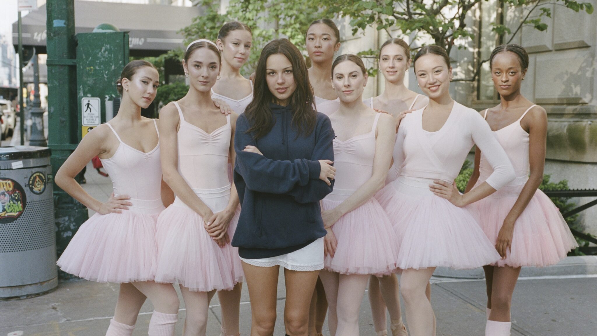 Olivia Rodrigo, a young woman wearing a large, blue, long-sleeved shirt, whort white skirt, and black knee-length boots, poses with her arms crossed with eight ballerinas. The dancers are all costumed in pink tutus. They pose outside on a New York City sidewalk.