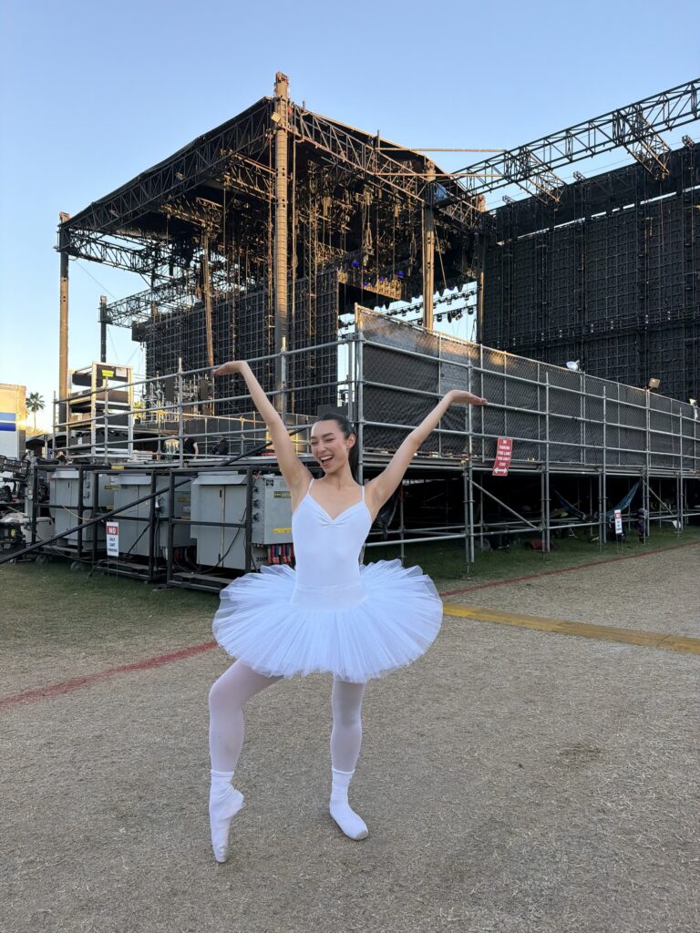 A young woman in a white tutu, white tights, and white socks over pointe shoes poses in back of a large outdoor stage. She pops her right foot up onto pointe and throws her arms up, palms flexed, and wears a large, enthusiastic smile.