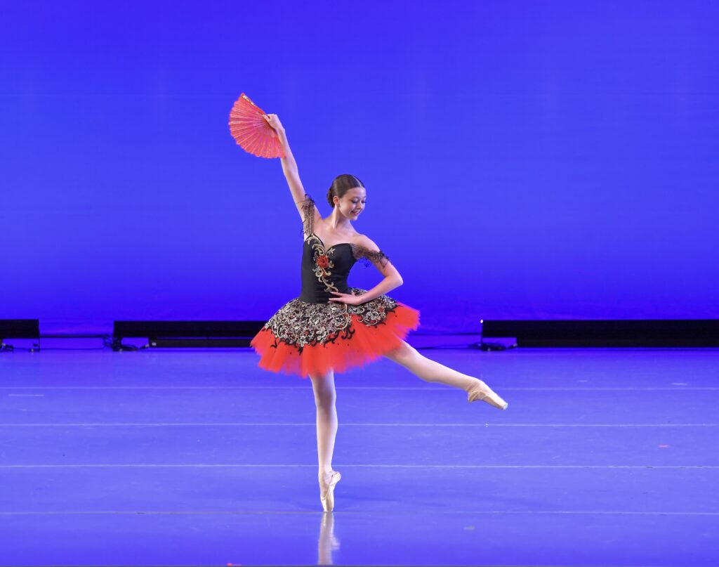 Evelyn Allen lifts one leg a la seconde while balancing en pointe, one hand on her hip and the other extended up. She wears a red, gold, and black tutu and holds a red fan.
