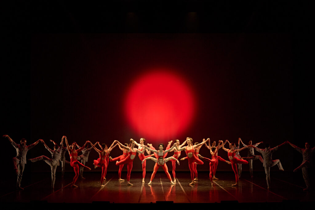 A large ensemble of dancers in red unitards and short tan dresses form a tableau onstage. The hold hands and lift their arms, lifting a leg in attitude devant or derriere. In the middle, one man spreads his legs and leans forward with his arms curved down as another dancer rests on his back, arching upward. A red circle is illuminated on the dark backdrop behind them.