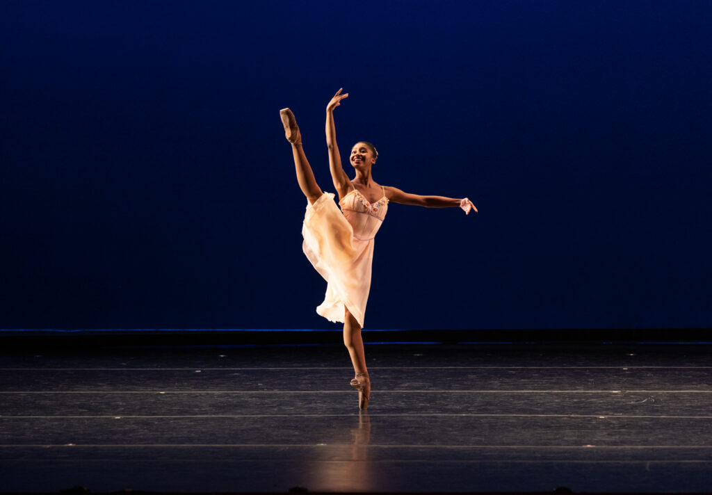 Onstage, Madison Brown lifts one leg energetically in a la seconde with her standing leg on pointe. She holds her arms in a high third as she looks toward her top hand. She wears a flowing peach-colored dress.