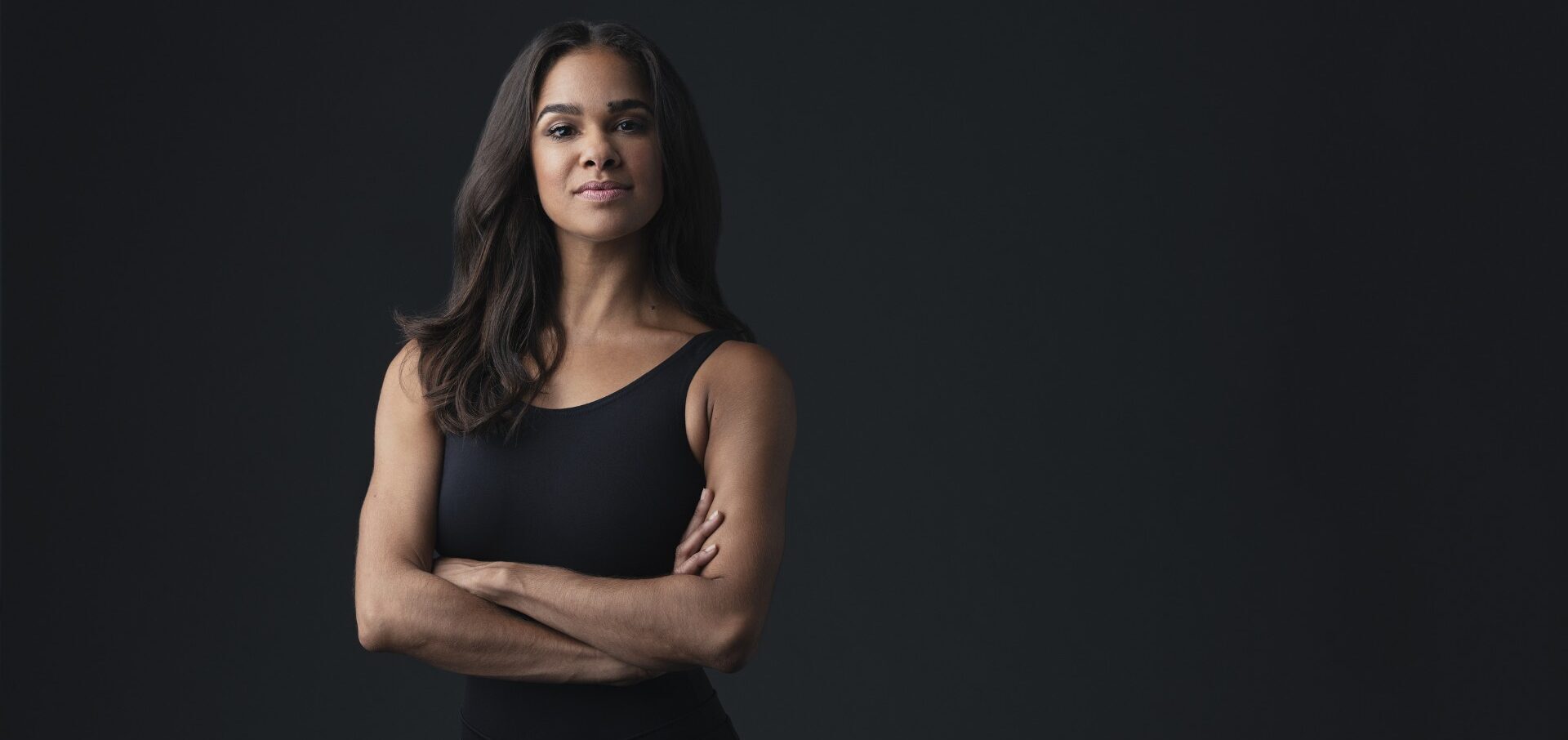 Misty Copeland stands with her arms crossed confidently and gives a sly smile as she poses for a portrait in front of a dark gray backdrop.