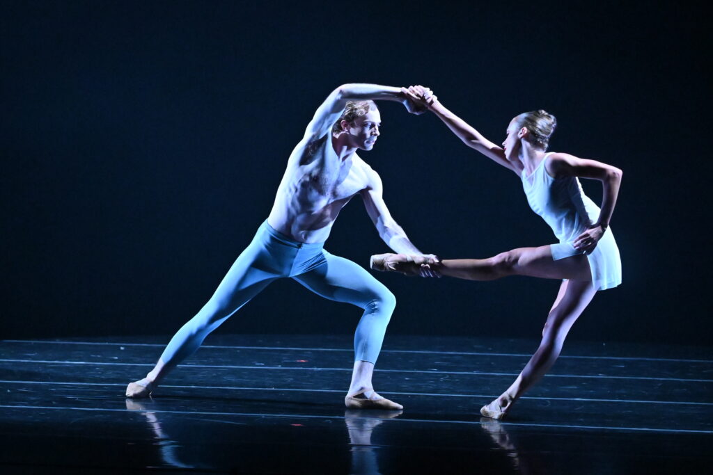 Onstage in dark blue lighting, two dancers perform a pas de deux. One dancer lunges forward while holding his partner's hand and foot. The other dancer leans backward with one leg lifted front, one arm bent with her hand at her hip, and the other arm reaching up to hold her partner's hand.