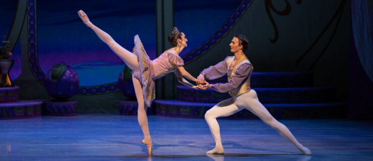 Onstage, two dancers in purple, white, and gold costumes perform a pas de deux. One dancer does a penché on pointe, holding her partner's hands in front of her, as he lunges in toward her.