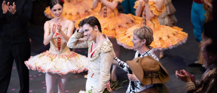 Onstage after a show, Skylar Brandt, Jake Roxander, and Susan Jaffe stand surrounded by corps dancers in yellow tutus as everyone applauds Roxander. Jaffe places one hand on Roxander's back as he runs a hand through his hair in happy disbelief, holding a bouquet of flowers in the other.