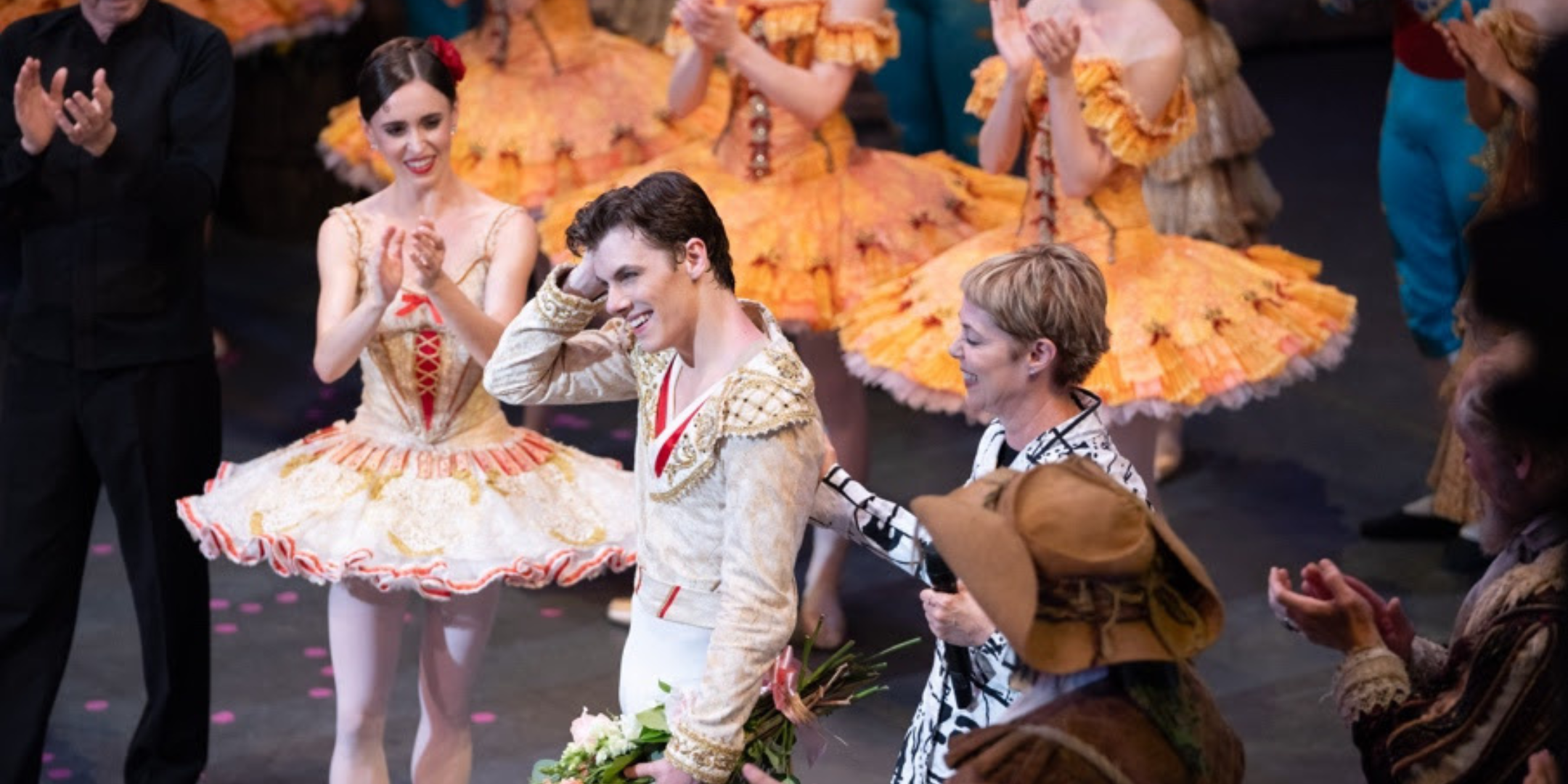 Onstage after a show, Skylar Brandt, Jake Roxander, and Susan Jaffe stand surrounded by corps dancers in yellow tutus as everyone applauds Roxander. Jaffe places one hand on Roxander's back as he runs a hand through his hair in happy disbelief, holding a bouquet of flowers in the other.