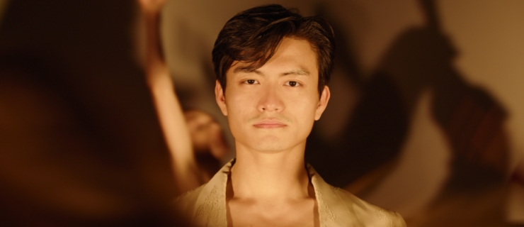 A close-up of Chun Wai Chan, obscured partially by another dancer in motion. Chan stands still with a soft light shining on his face, and he peers into the camera lens with a pensive expression. Shadows of other dancers project onto the wall behind him.