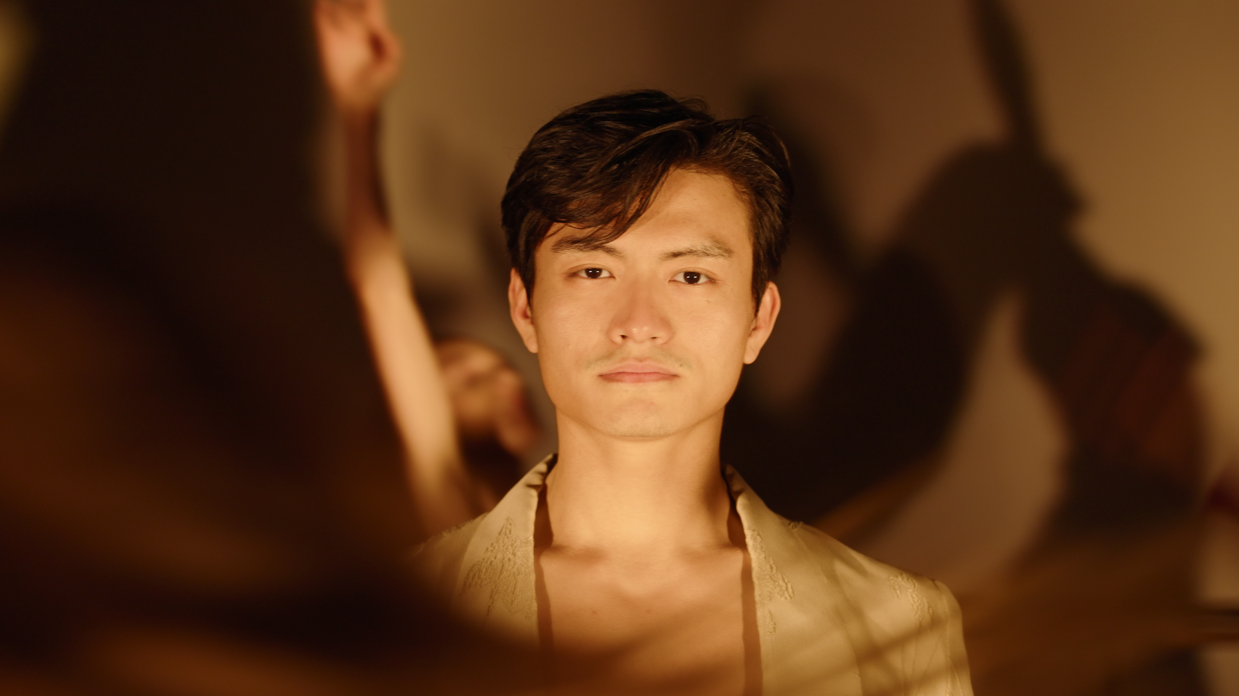A close-up of Chun Wai Chan, obscured partially by another dancer in motion. Chan stands still with a soft light shining on his face, and he peers into the camera lens with a pensive expression. Shadows of other dancers project onto the wall behind him.
