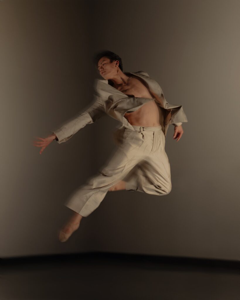Chun Wai Chan, partially blurry for an action shot, jumps with one leg tucked in retiré derriere as he leans sideways toward his bottom leg, which is extended on the diagonal downstage left. He reaches with his left arm and looks in that direction as his other arm lifts softly. He wears a light tan suit jacket and slacks, with the jacket open to expose his bare chest.