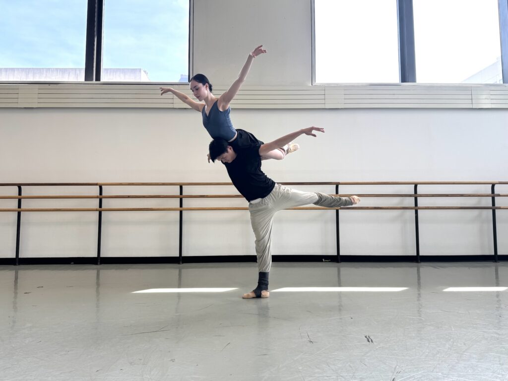 Chun Wai Chan, with one leg lifted in arabesque, balances Mira Nadon in a "Bluebird" lift across his shoulders. He tilts his head down and lifts his arms to either side as Nadon arches up with her hips on the backs of his shoulders and neck. She bends her legs slightly, ankles crossed, and lifts her arms up gently as she seems to hover above him. They rehearse in a large ballet studio and wear practice clothes.