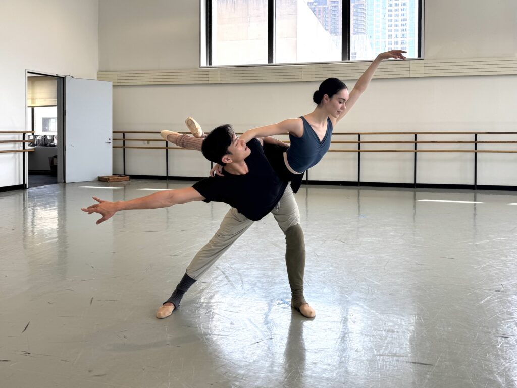 In a large ballet studio, Chun Wai Chan lunges sideways as he holds Mira Nadon's waist to balance her on the back of his hip. Nadon crosses her ankles and lifts one arm up, her eyes looking down and out, as she wraps her other arm around Chan's shoulders. Chan looks at her and extends his other arm to the side.