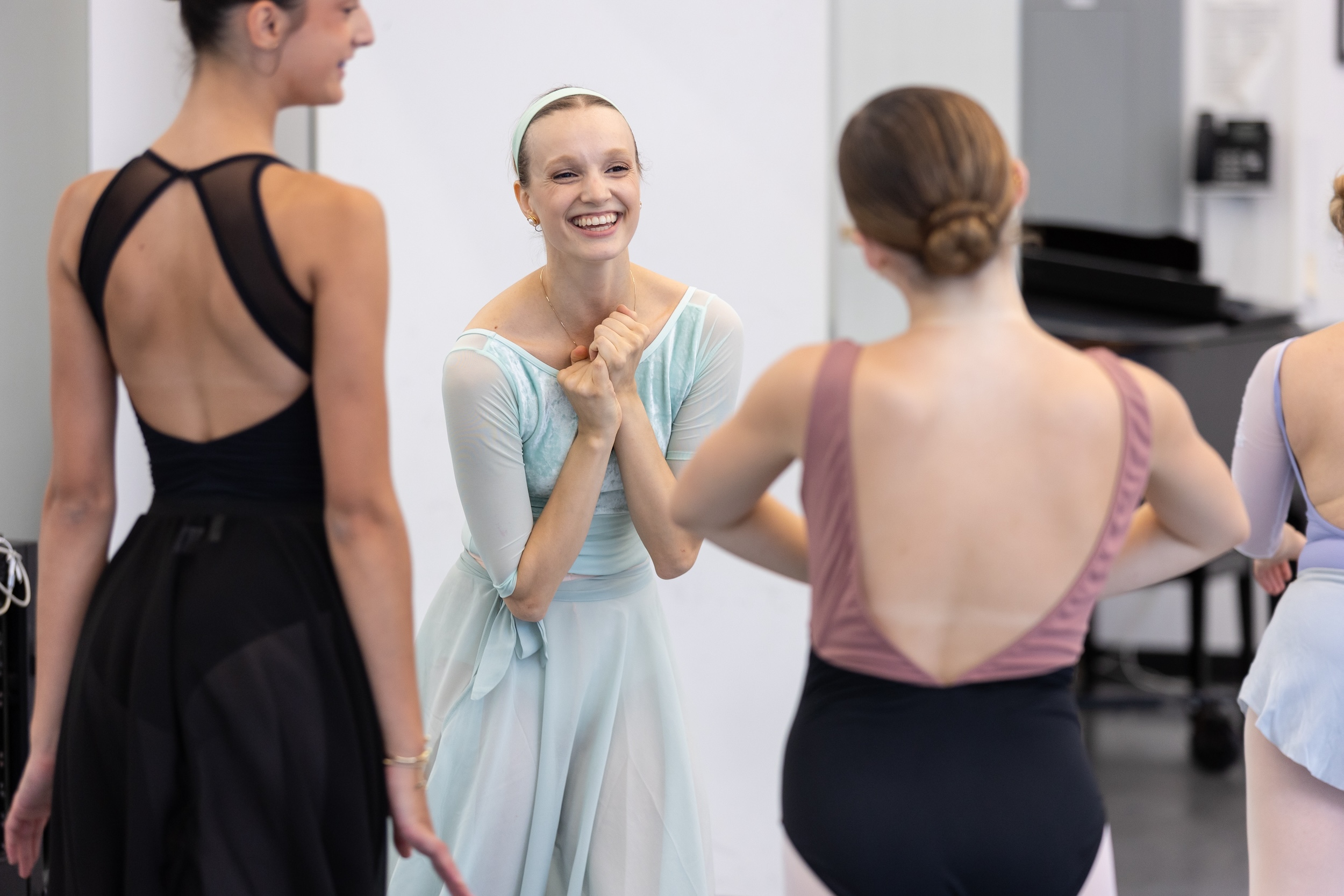 A female ballet teacher wearing a int green leotard and matching skirt and headband clasps her hands to her chest and smiles warmly to a group of teenage ballet students.