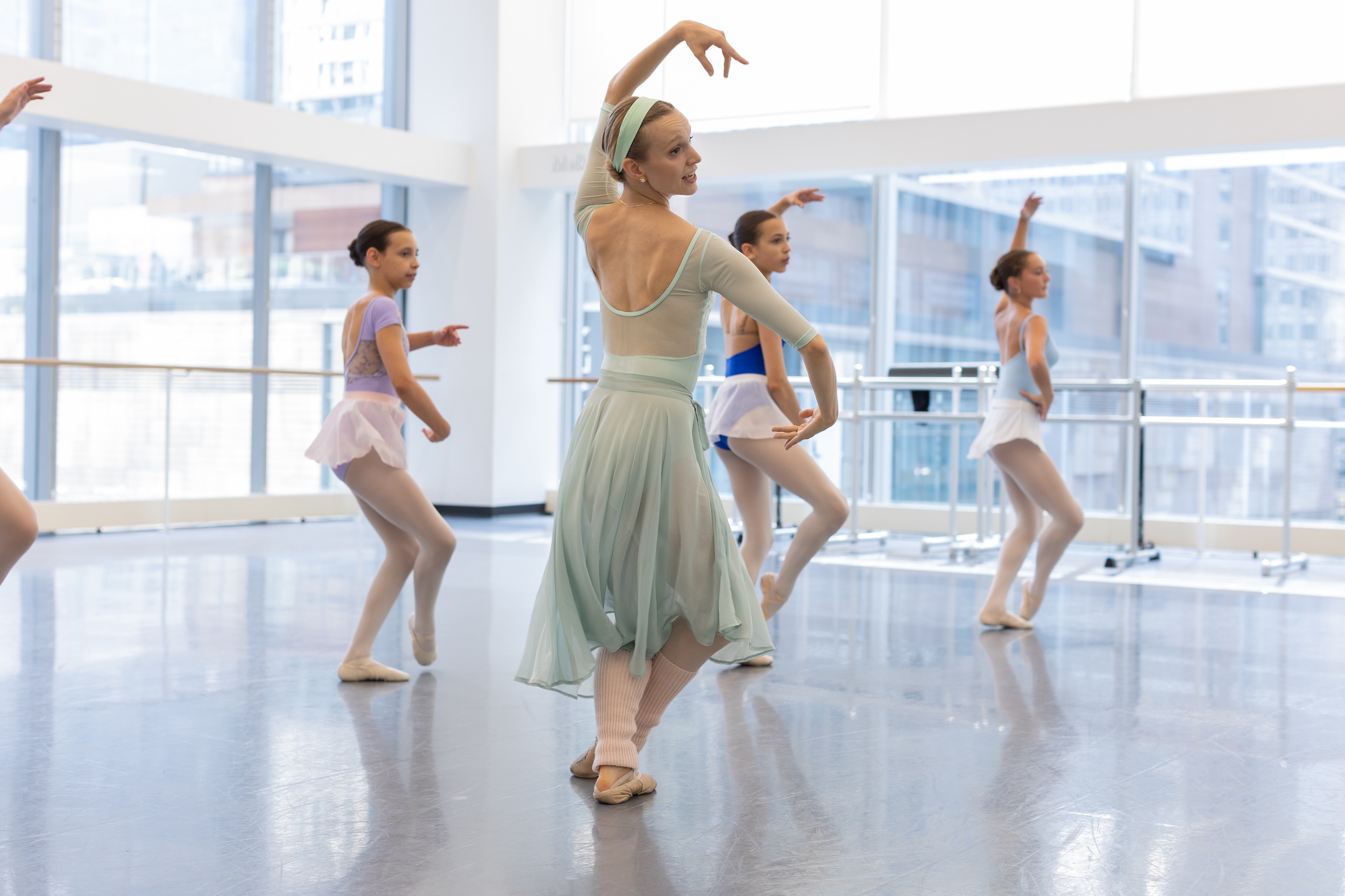 A female ballet teacher wearing a mint green leotard and matching skirt, pink tights and ballet slippers, and pink leg warmers, demonstrates a step during a repertoire class in a sunny dance studio. A group of young ballet students copy her moves.
