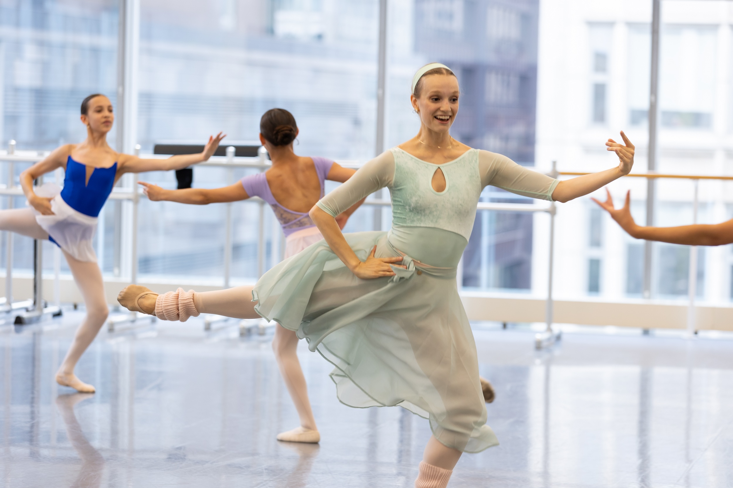 A female ballet teacher wearing a mint green leotard and matching skirt, pink tights and ballet slippers, and pink leg warmers, demonstrates a sissone during a repertoire class in a sunny dance studio. A group of young ballet students copy her moves.
