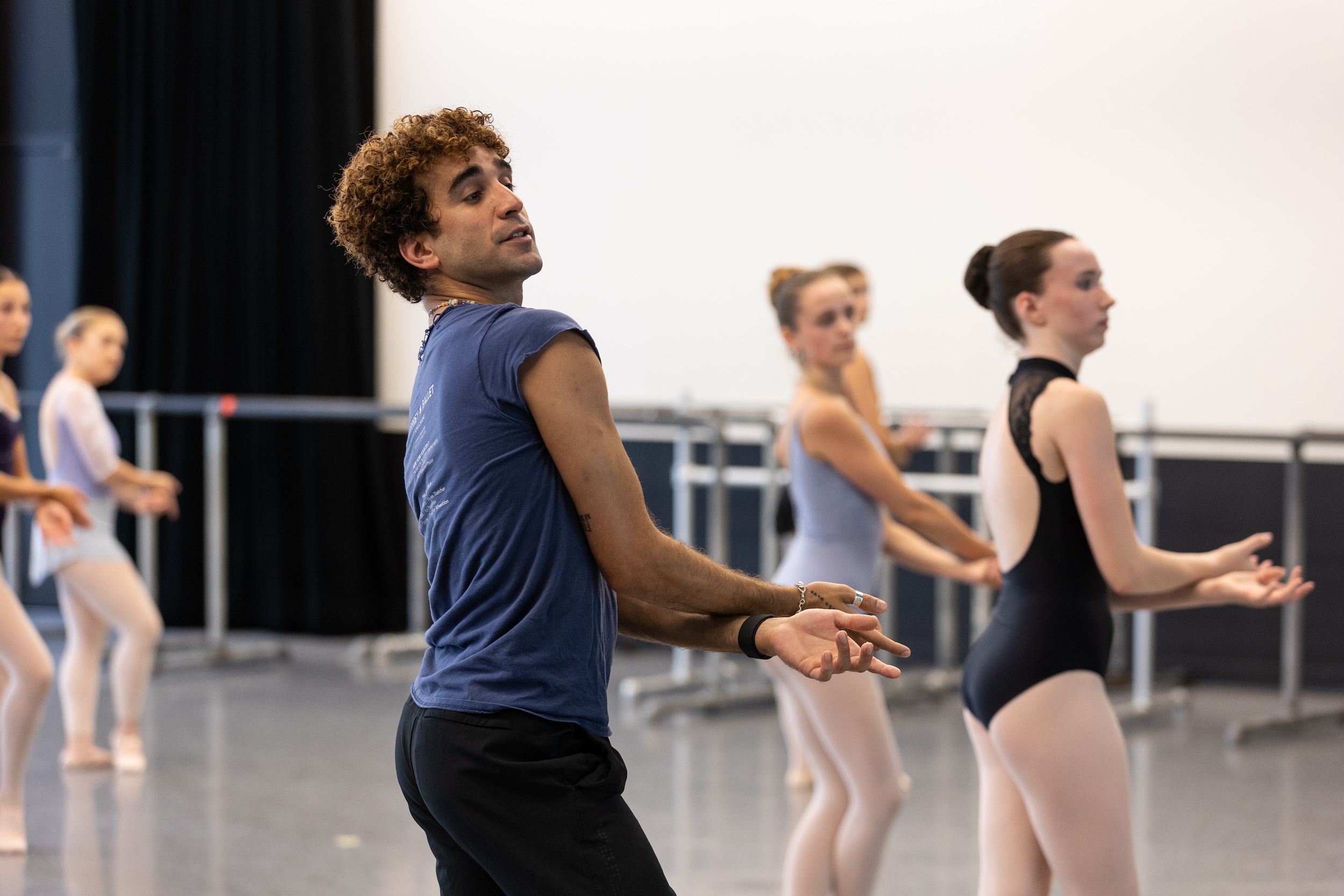 A male ballet teacher in a blue t-shirt and black pants crosses his wrists and twists his body slightly, demonstrating a dance. His teenage students, wearing various leotards and tights, follow his movements in the background.