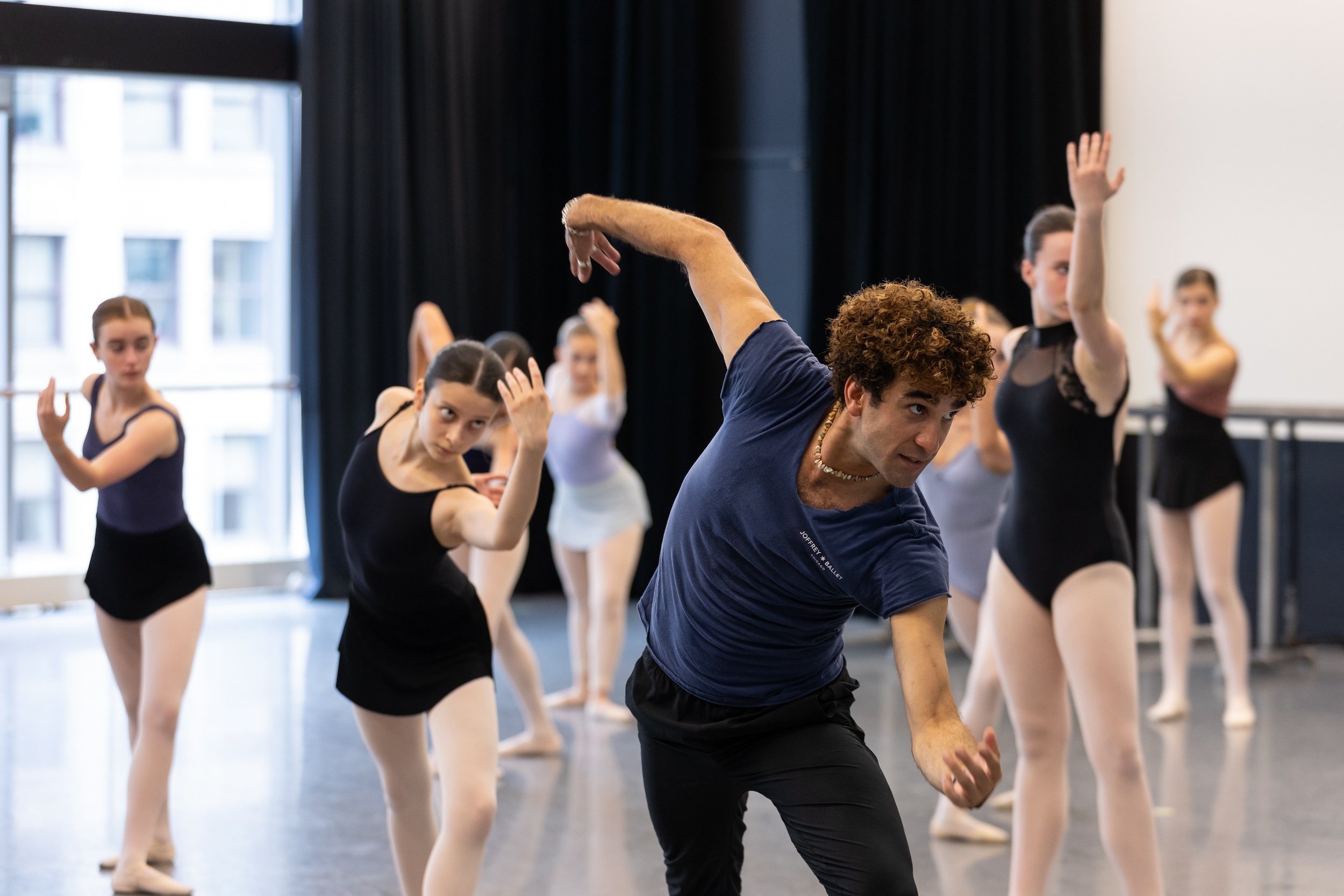 A male ballet teacher in a blue thirt and black pants tilts his body, his arms curved, as he demonstrates choreography. His teenage students, wearing various leotards and tights, follow his movements in the background.