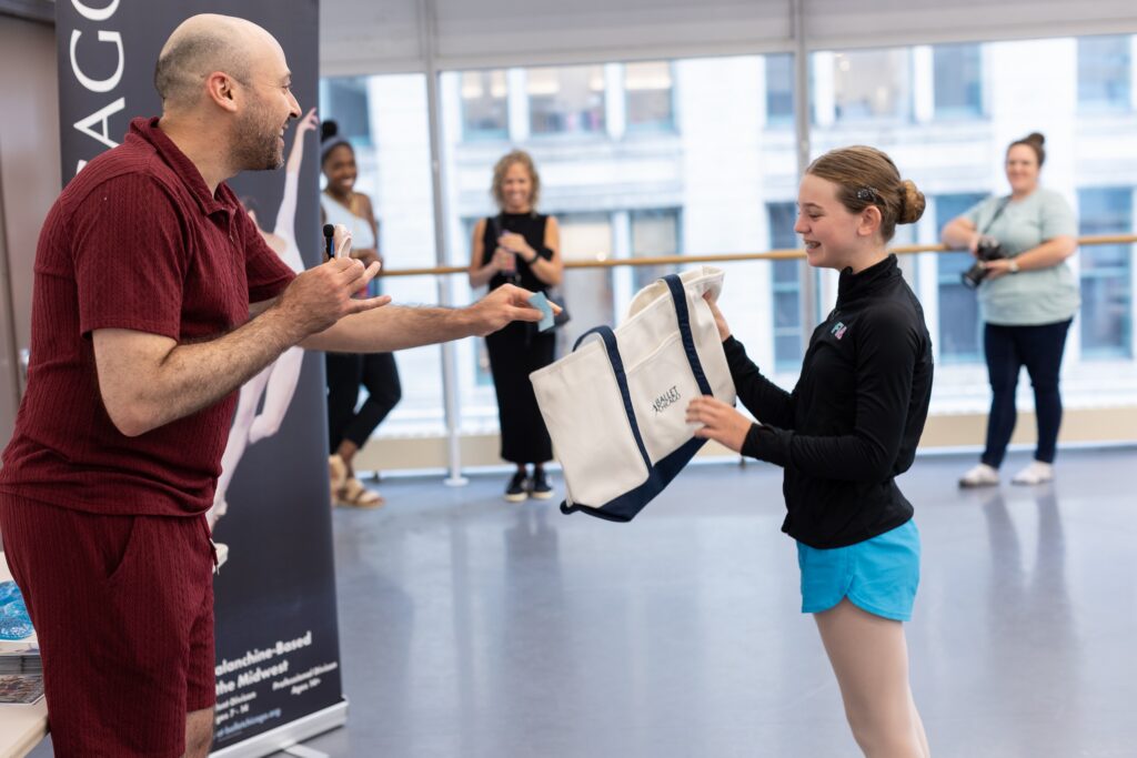 A man wearing a burgundy shirt and shorts hands a tote bag to a teenage ballet student, who wears a black zipup turtleneck, blue shorts, and pink tights.