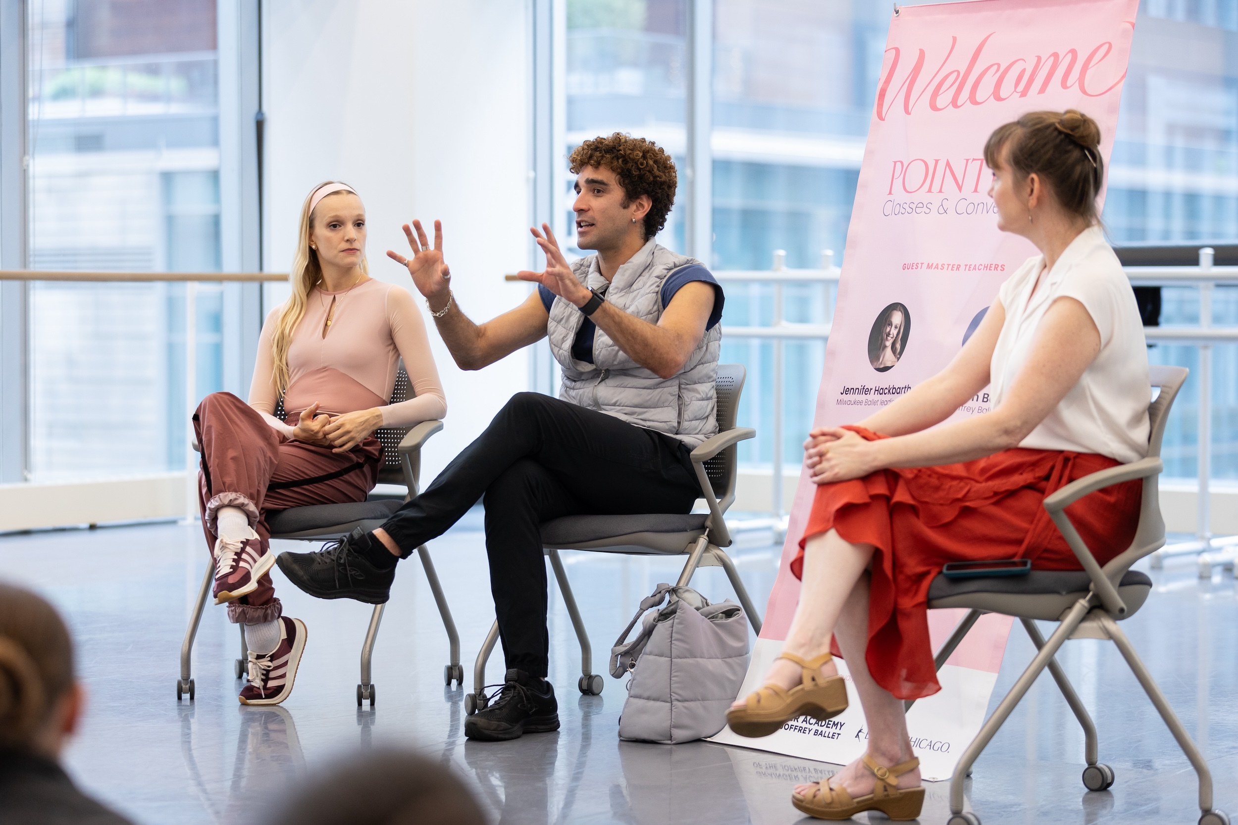 Three adults sit in chairs in a large ballet studio and talk to a audience (out of view). A pink banner is in the middle that says "Welcome to Pointe Classes & Conversations."