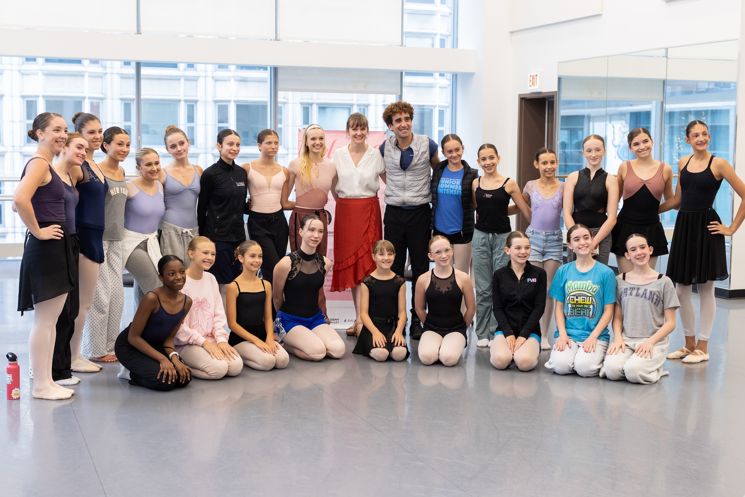 A group of ballet students and teachers pose for a large group photo in a large dance studio with floor-to-ceiling windows.