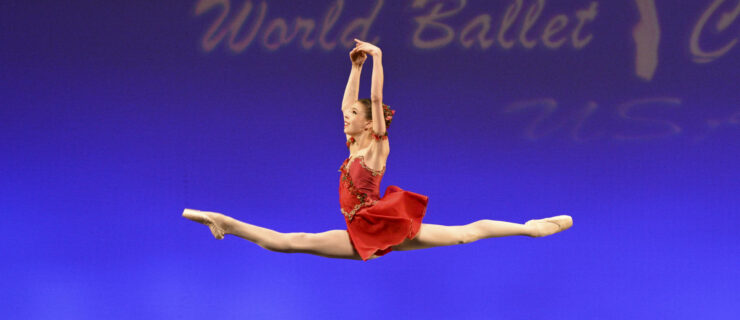 Megan Jarvis flies in a saut de chat with her arms in high fifth. She wears a shirt red dress and tiny red flowers around her bun.