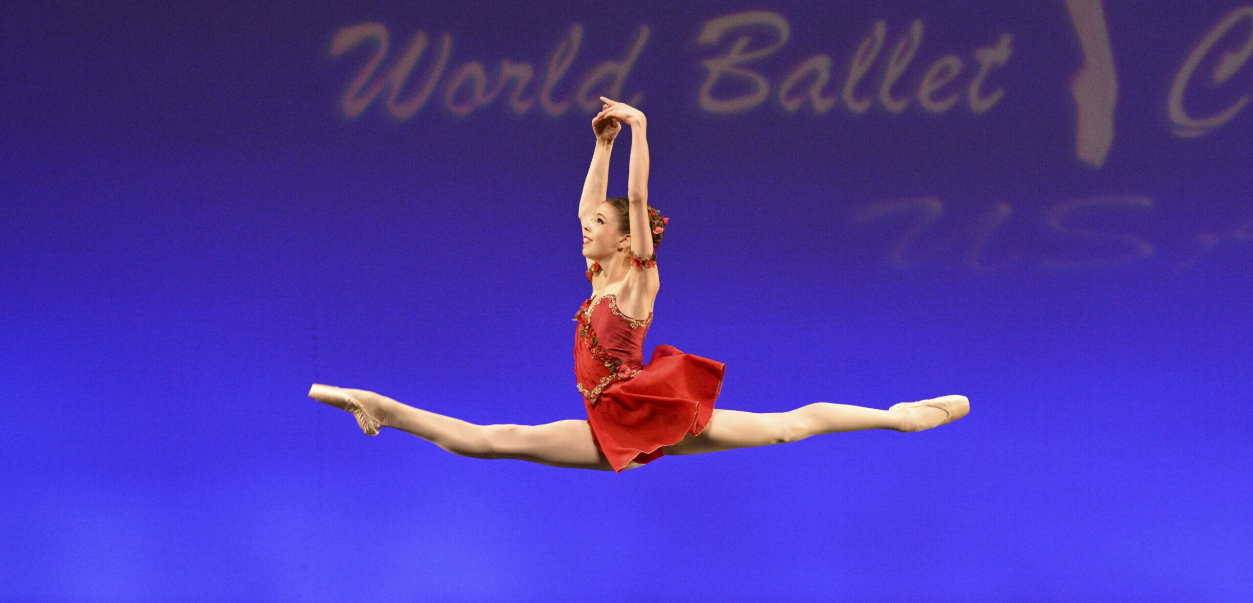 Megan Jarvis flies in a saut de chat with her arms in high fifth. She wears a shirt red dress and tiny red flowers around her bun.