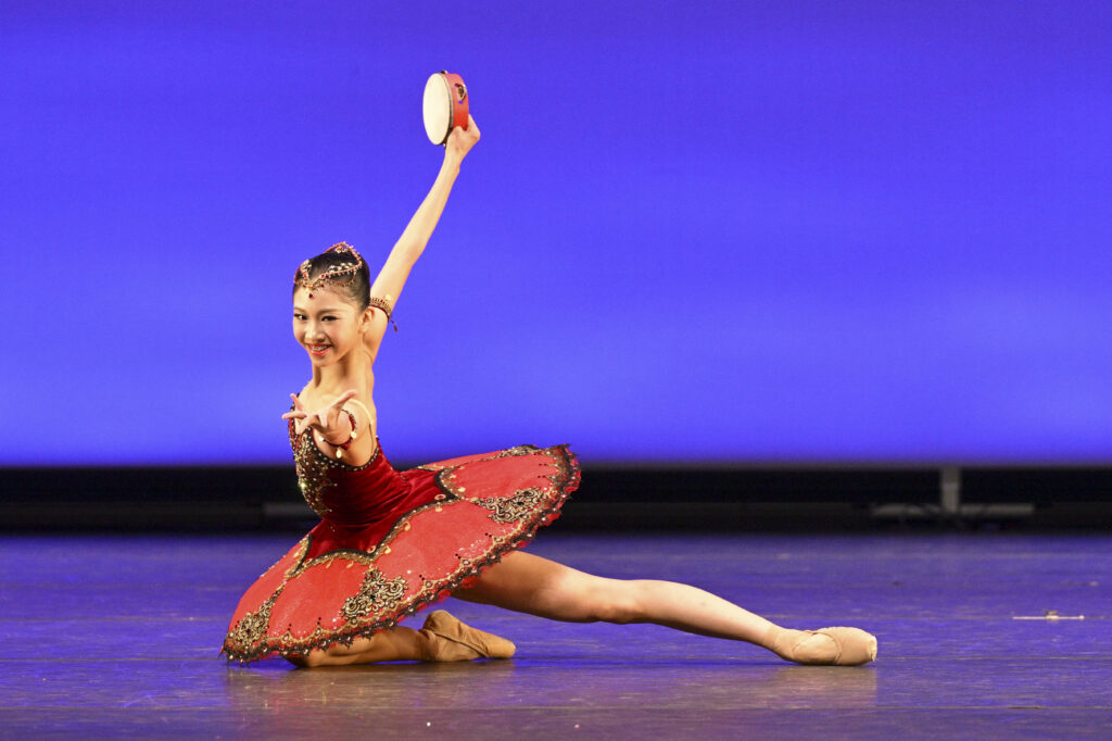 Elynn Nie tucks one leg under and extends the other to the side as she twists her torso, reaching front with one arm and back and up with the other. She wears a black and red tutu and holds a tambourine in her back hand as she grins at the audience daringly.