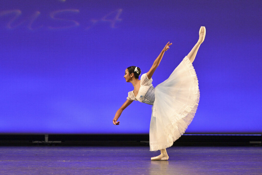 Minori De Silva penchés forward with her bottom leg in fondu and her arms in third. Her back arm matches the line of her arabesque leg. She wears a light blue and white peasant dress costume.